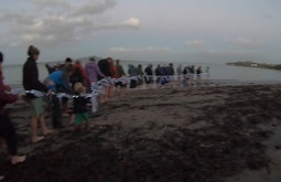 Dozens of people stand in a line on a beach at dusk holding a rope with lights.