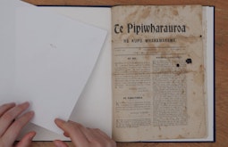 A birds-eye view of someone's hands turning pages in a book. The heading of the book reads "Te Pipiwharauroa"