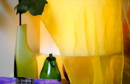A yellow piece of fabric is draped over a lamp. There is a green vase with a rose beside the lamp.