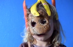 A child sits wearing carrots attached as ears, a banana on their head, and slice of bread with eye holes cut out.