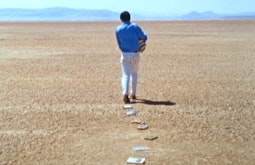 A person walks in a desert carrying an armful of books, some are strewn behind them in a line.