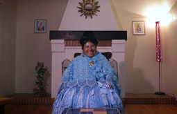 A Bolivian woman dressed all in light blue is seated in front of a fireplace facing the camera, two paintings of Jesus are hung on the wall behind her.