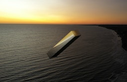 A digitally rendered metal oblong flies above the sea while the sun sets.
