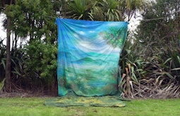 A large piece of fabric painted with an image of islands and trees hangs from a cord suspended amongst native New Zealand bush.