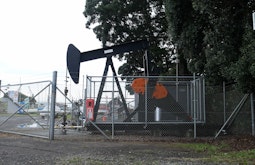 An oil well dredges behind large metal fences with warning signs attached. Parked cars are in the background.