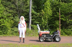 A man wearing red and white robes stands on a driveway, behind him are tall green trees. A ride on lawnmower with several decorations are attached to it.