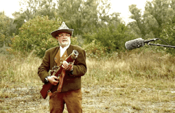 A man wearing a wool suit and pointed hat stands in a grassy field holding a large antique rifle. A microphone protrudes into the frame from the right.