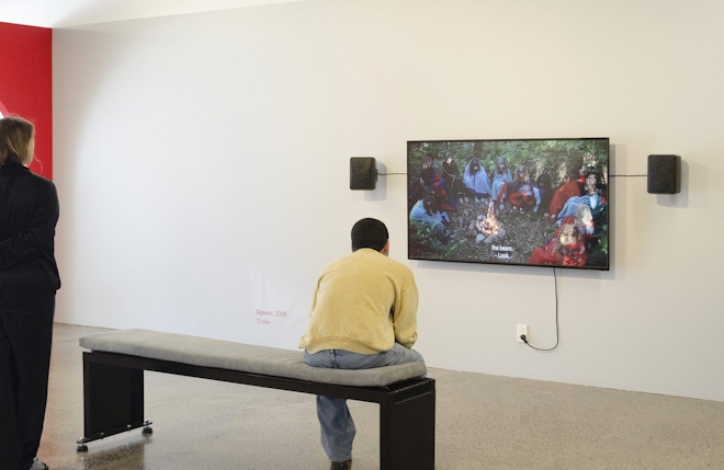 A man sits on bench and watches a film on a wall-based TV.