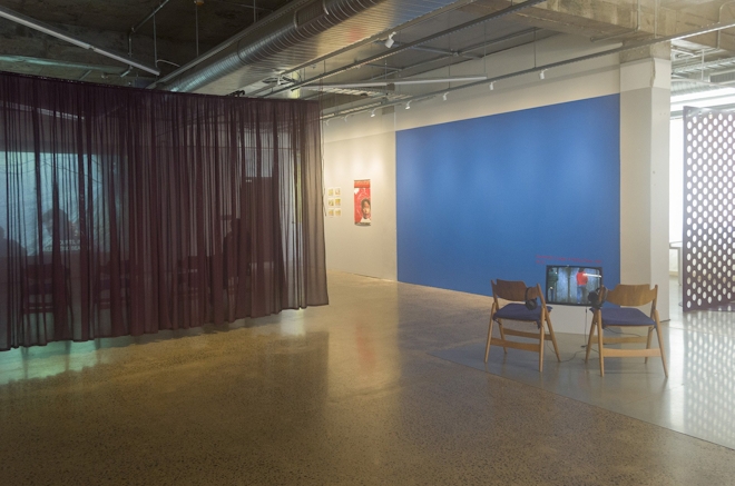 A gallery space is bisected by a sheer curtain; two chairs face a TV monitor; one wall is bright blue