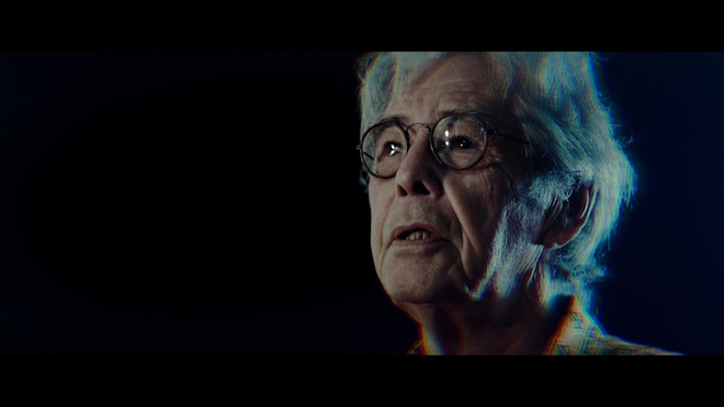 An older person wearing glasses speaks to an unseen interviewer. Their hair is tinged with psychedelic blue light.