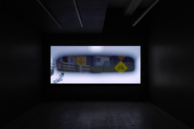 A film plays in a dark screening room; the screen depicts a view of a car's rear vision mirror showing various rural street signs