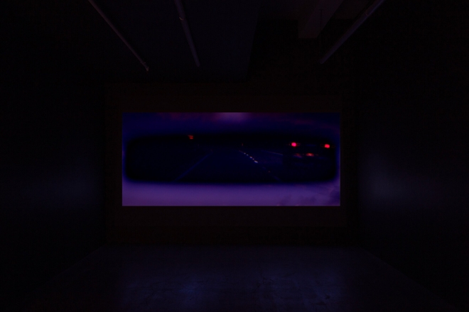 A film plays in a dark screening room; the screen depicts a view of a car's rear vision mirror showing a passing car's headlights. The view is very dark.
