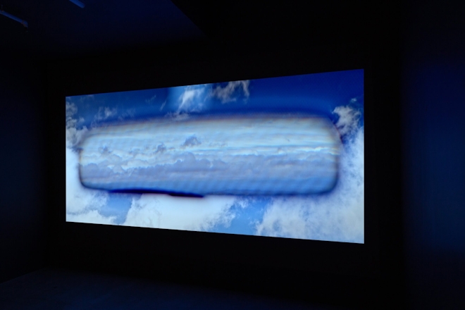 A film plays in a dark screening room; the screen depicts a view of a car's rear vision mirror showing a cloudy sky; the clouds also fill the view out the front window of the car for a total sky effect