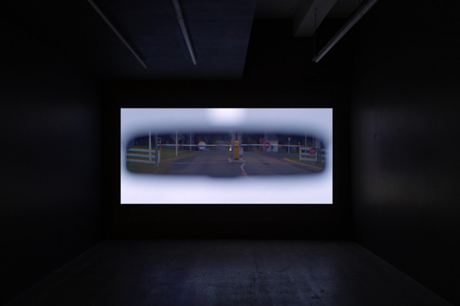 A film plays in a dark screening room; the screen depicts a view of a car's rear vision mirror showing a railway crossing