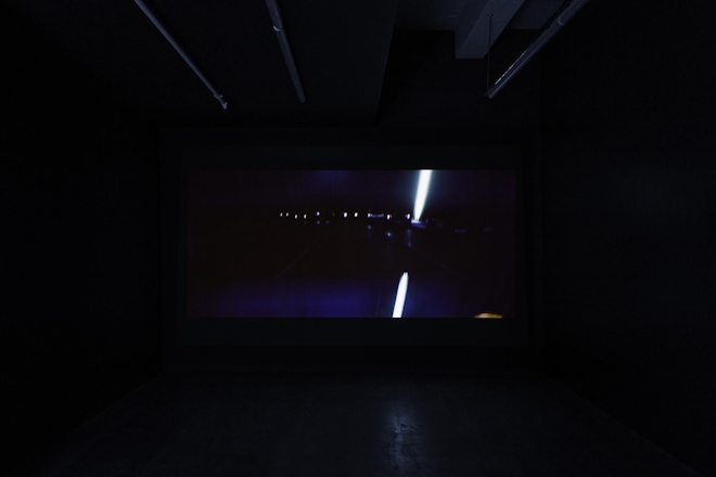 A film plays in a dark screening room; the screen depicts a view of a car's rear vision mirror showing road lights at night. The view is very dark.