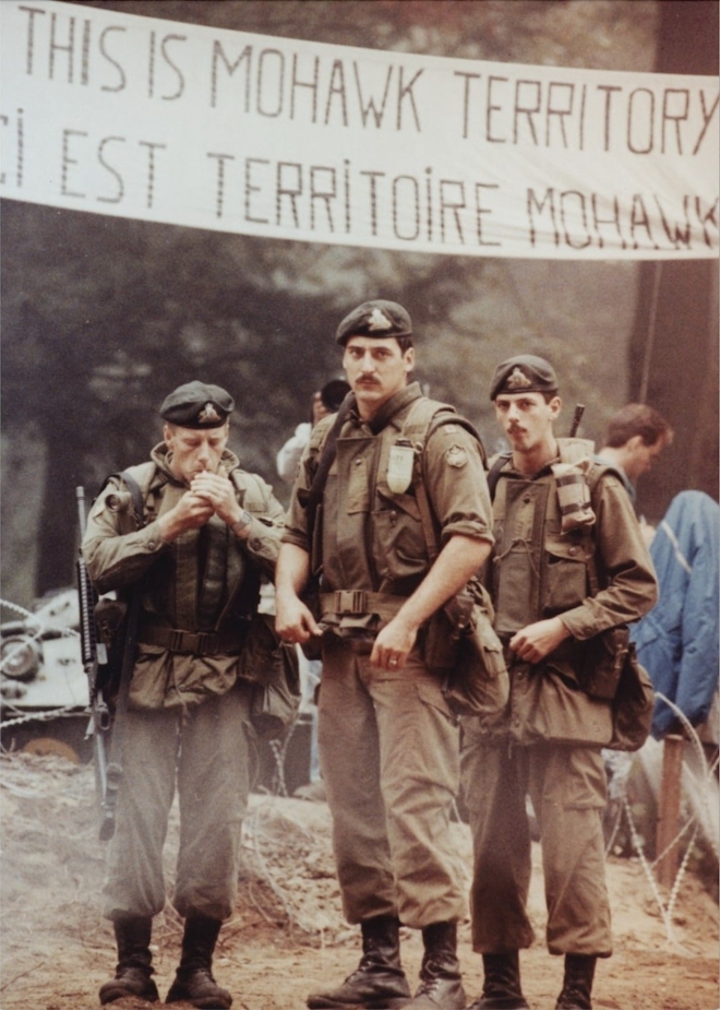 Three men in military uniform stand in a forest underneath a banner that reads 'This is Mohawk Territory'