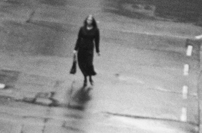 A black and white film still of a young woman walking in a deserted city street