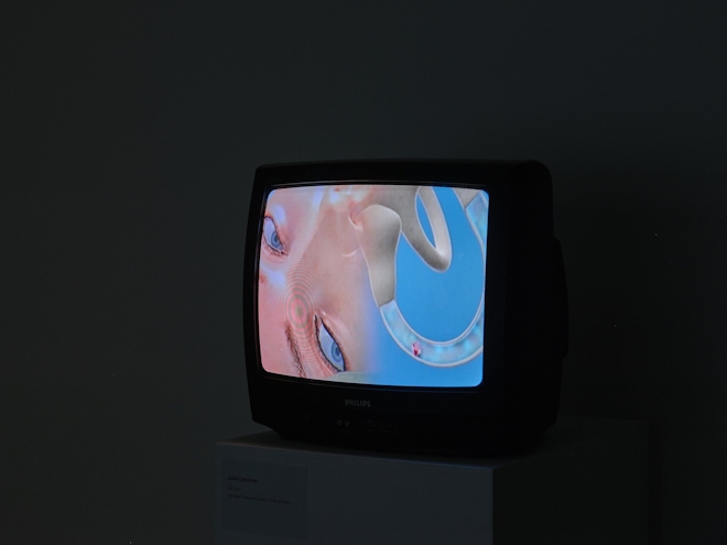 A 1990s TV monitor shows an animated image of a woman's upper face, and a tube which is arching towards her nose. In the transparent tube is a small red object.