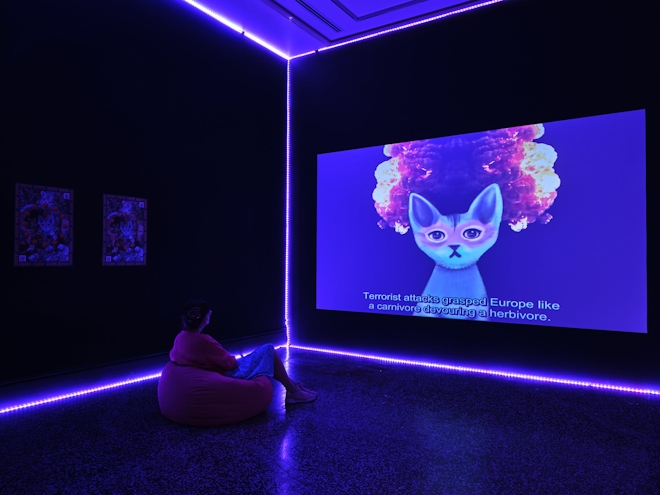 A person in a dark room is slightly illuminated by a large projection screen playing a video. The space is also illuminated by a LED lighting strip which is outlining the corners of the room. A person sits on a beanbag enjoying the video installation