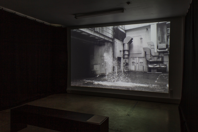 Installation view of a black and white film projected in a darkened room. The image on screen shows an explosion of feathers in an alley-way.