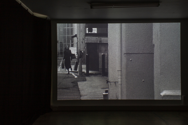 Installation view of a black and white film projected in a darkened room. The image on screen depicts a young woman walking in an alley-way.