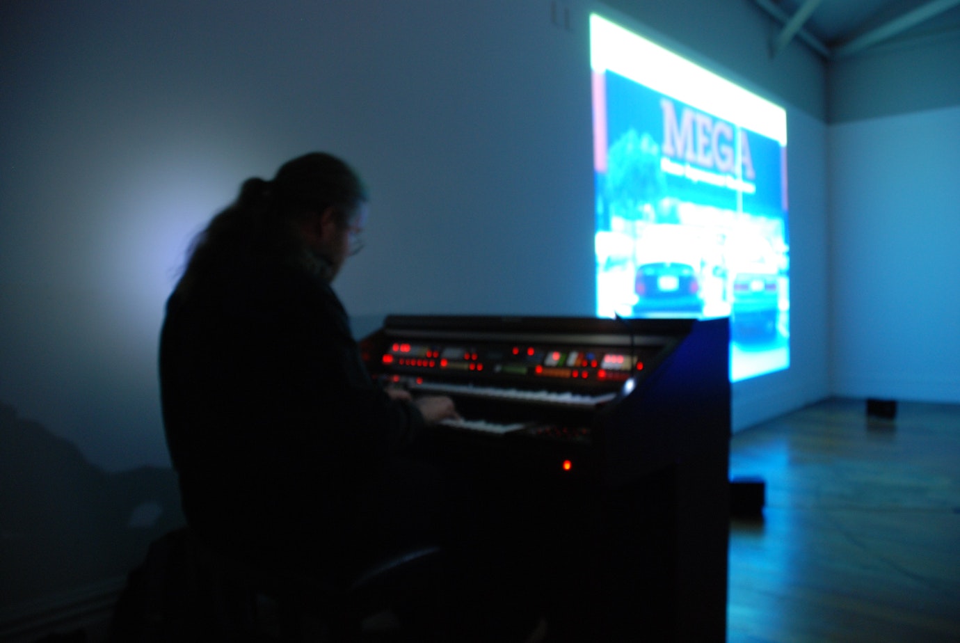 In the dark of a gallery a person plays a keyboard organ to accompany a projected image of a Mitre 10 mega store