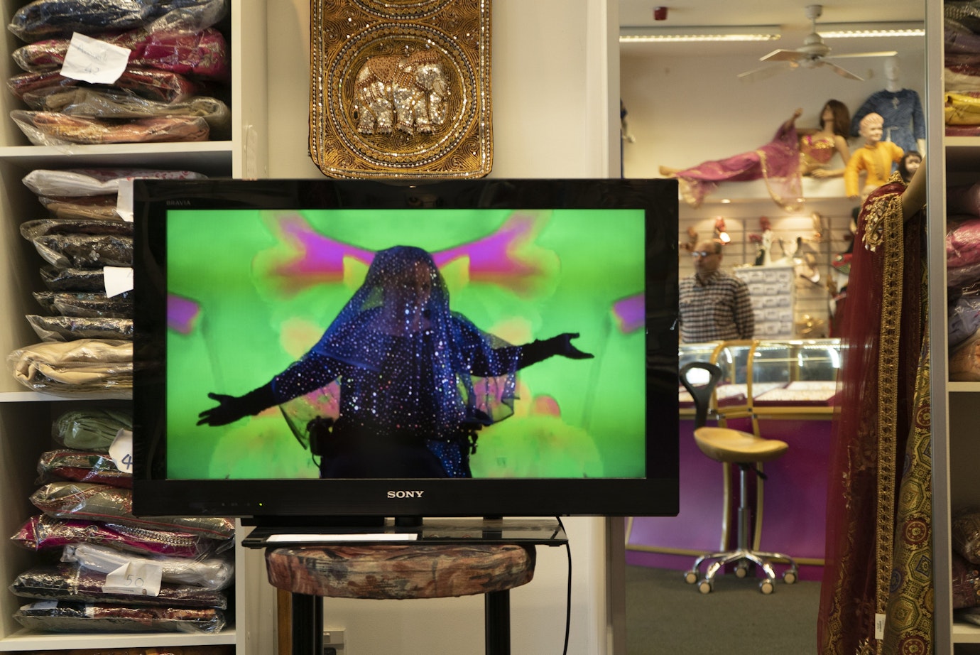 An installation of a video artwork on a monitor in a shop. The shops sells colurful Indian fashion. The woman in the video dances in brightly coloured clothing.