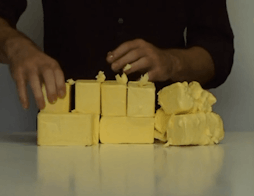 Yellow blocks of butter are stacked and formed on a table by the artist who stands behind it