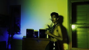 A person wearing a leather harness stands in a living room bathed in blue and green light looking at the camera.