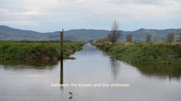 A calm river flows amongst grassy farmland. There are moutnains in the background and a fish jumping in the foreground. Subtitles read "between the known and unknown"