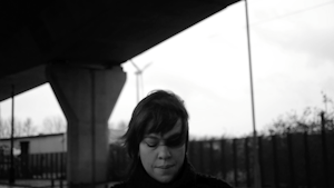 A black and white image of a person underneath a road, motorway or overpass. The person appears looking down with their dark short hair blowing in the wind, slightly obscuring their face. The background is blurred and the image has a soft feel.