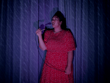 Ash Ulutupu wears a red dress sings alone into a microphone  in front of a blue stage curtain