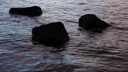Three shadowy rocks sit up out of the bright metallic-like ocean, water swirls creating patterns around the rocks