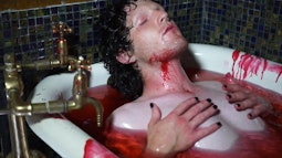A person lies in a bath holding their chest, the water is red with fake blood. There is blood running down their neck and the side of the bath.