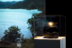 Inside a museum vitrine is a black motorbike helmet with a large video projection behind