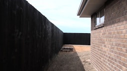 A brick house is surrounded by a tall black wooden fence, the ground is dry and bare.