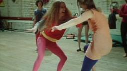 In a dance studio 2 people wearing pink outfits dance with each other.