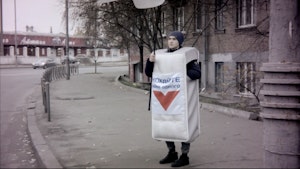 A person stands a footpath waving a large advertisement sign wearing a mattress costume.