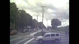 At an intersection cars wait, people walk by. A large row of trees runs beside the road.