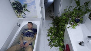 A still from Alex Plumb’s work shows a person in the bath, fully clothed with a blank expression on their face. The image is taken from a high angle, looking down into the entire bathroom and down the hallway of the house. A large overgrown plant spills messily through the bathroom and down the corridor. The image feels airy, suspenseful, uncanny.