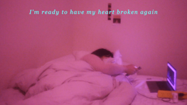 Natasha plays on their phone whilst watching tv off a laptop in bed in a pink room, text overlays the image reading "I'm ready to haver my heart broken again"
