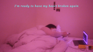 Natasha plays on their phone whilst watching tv off a laptop in bed in a pink room, text overlays the image reading "I'm ready to haver my heart broken again"