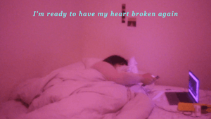 Natasha plays on their phone whilst watching tv off a laptop in bed in a pink room, text overlays the image reading "I'm ready to haver my heart broken again"