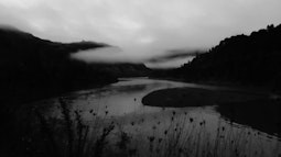 A black and white scene of a river running between dark hills. Low cloud hangs over the river.