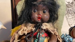 A doll with large black eyes and red lips wears customary Māori attire, there is a tiki hanging from the dolls neck.