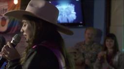 A woman wearing a cowboy hat sings into a microphone. Two audience members sit watching her in the background.