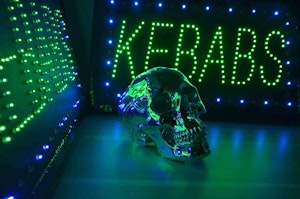 A mirror-like metalic skull sits between two neon signs that read "KEBABS"