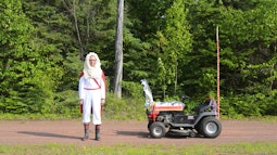 A man wearing red and white robes stands on a driveway, behind him are tall green trees. A ride on lawnmower with several decorations are attached to it.