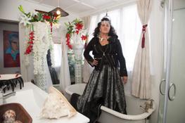 Suzanne stands up in her bathtub wearing a silky long dress, black jacket and traditional necklace and headpiece. She presents as powerful, serious, cool and confident