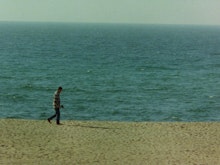 A man in a check shirt and jeans walks along a sandy beach holding a metal detector in front of him.